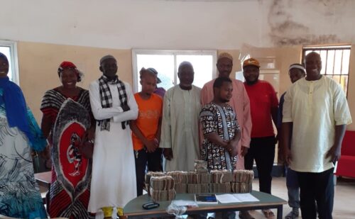 En gruppe af de lokale købmænd fotograferet på den dag, hvor de kunne hente betalingen for de leverede madvarer (skoleuniformer). På bordet ses bundter af gambianske pengesedler.